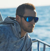 Man wearing sunglasses with a blue reflection, sitting on a boat with water in the background